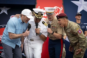 Secretary Pete Hegseth and three service members in uniform yell into microphones in front of a red, white and blue display outdoors.