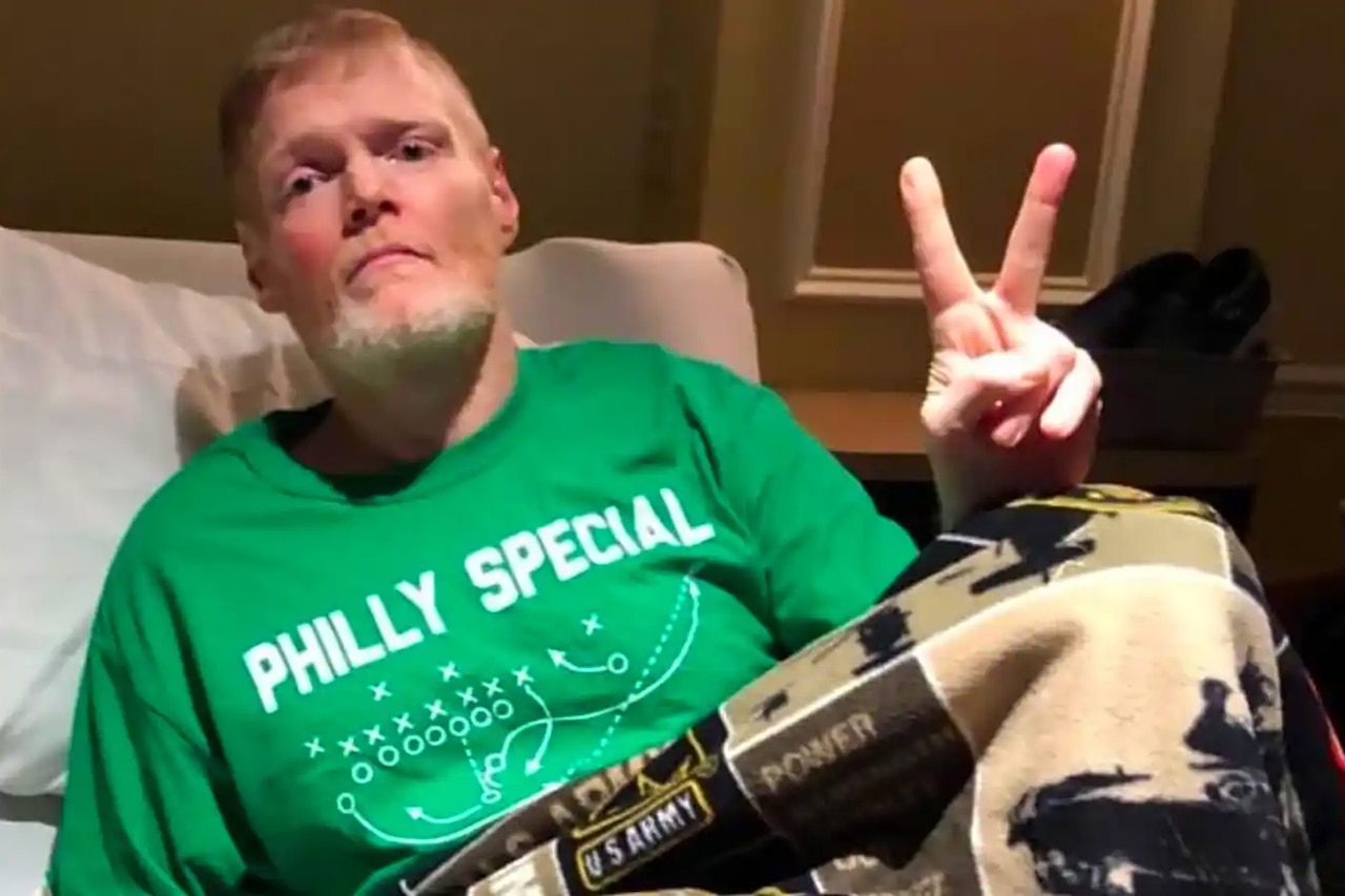 A man wearing casual attire sits in bed and flashes a peace sign.