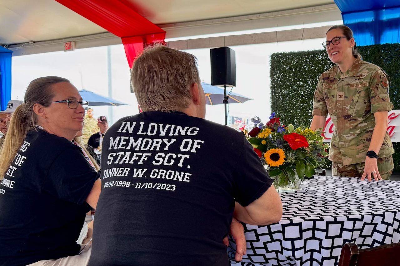 Two people sit at a table wearing black T-shirts that say, In loving memory of Staff Sgt. Tanner W. Grone, 08/18/19898-11/10/2023. A smiling woman wearing a military uniform is across from them.