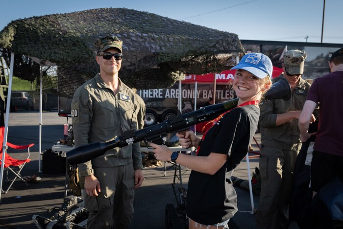A woman wearing casual attire and a blue cap poses with a weapon while a man, wearing a military uniform, smiles in the background.