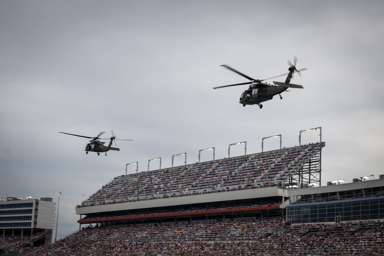 Two military helicopters fly above a stadium filled with people.