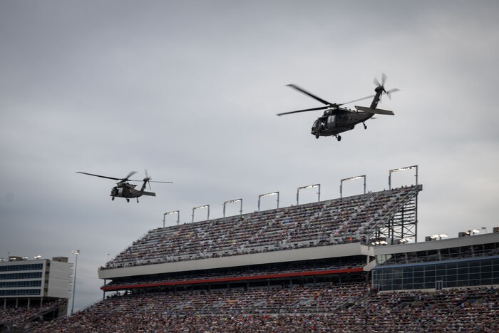 Two military helicopters fly above a stadium filled with people.