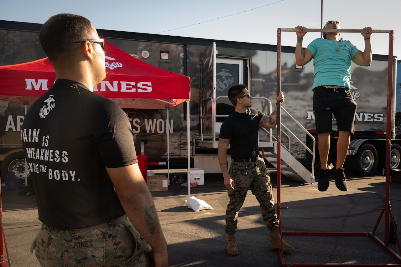 Two men in military uniform pants and Marine T-shirts stand next to a pullup device as another man wearing casual attire does pullups.