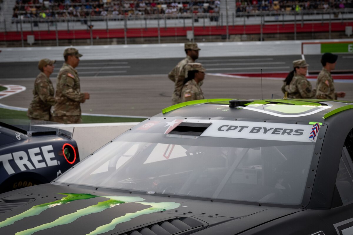 Six people wearing military uniforms are running past two race cars, which are parked on a racetrack. One of the cars has a sign that says "CPT Byers" on the windshield.