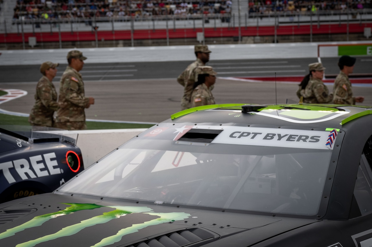 Six people wearing military uniforms are running past two race cars, which are parked on a racetrack. One of the cars has a sign that says "CPT Byers" on the windshield.