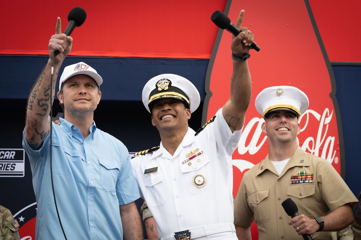 Three men with microphones look up at the sky. One is wearing casual attire, and the other two are wearing military uniforms.