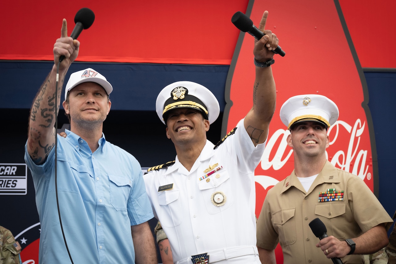 Three men with microphones look up at the sky. One is wearing casual attire, and the other two are wearing military uniforms.