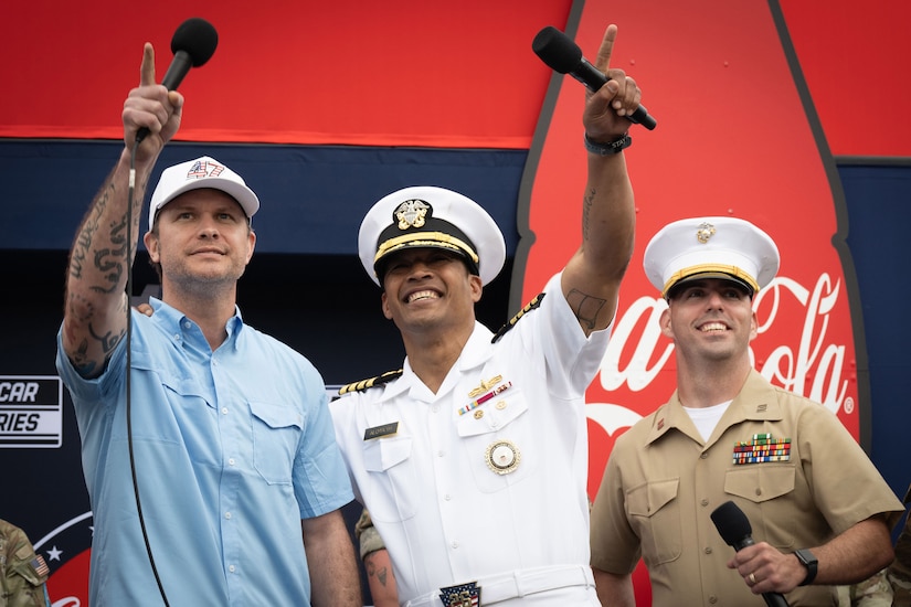 Three men with microphones look up at the sky. One is wearing casual attire, and the other two are wearing military uniforms.