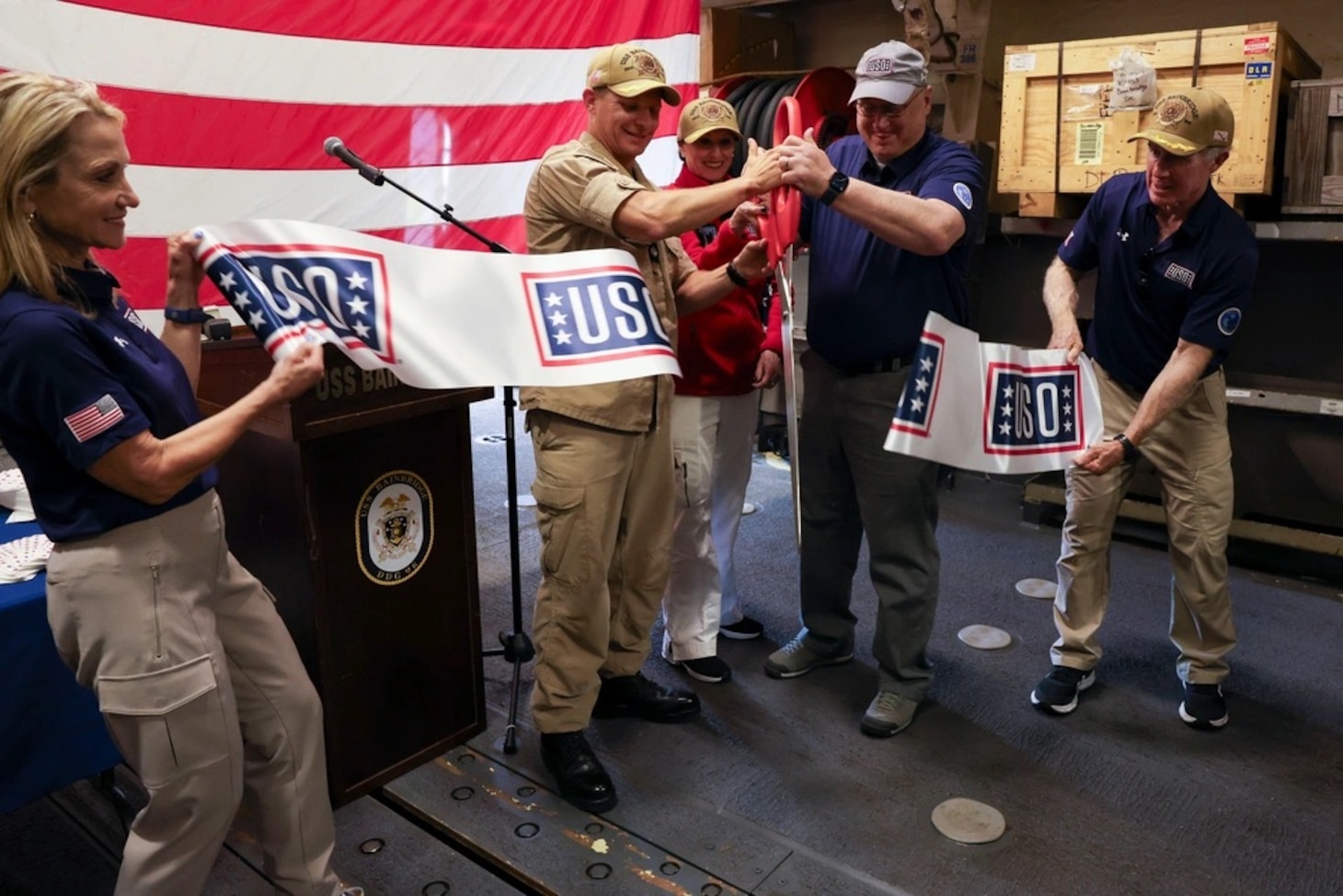 Cmdr. Raymond Miller IV, center left, commanding officer of Arleigh Burke-class guided-missile destroyer USS Bainbridge (DDG 96), and Mr. Christopher Plamp, Chief Operating Officer, United Service Organizations, cut a ribbon in the hangar bay in honor of the ship’s new USO unstaffed entertainment center, May 22, 2025. Bainbridge is a part of Carrier Strike Group 12, which provides combatant commanders and America’s civilian leaders multi-domain power to deter adversaries, underpin American security and economic prosperity, and project global power. (U.S. Navy photo by Mass Communication Specialist Seaman Cesar Licona)