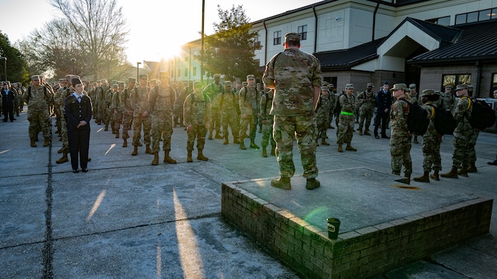 A man in military uniform stands in front of Air Force trainees outside.