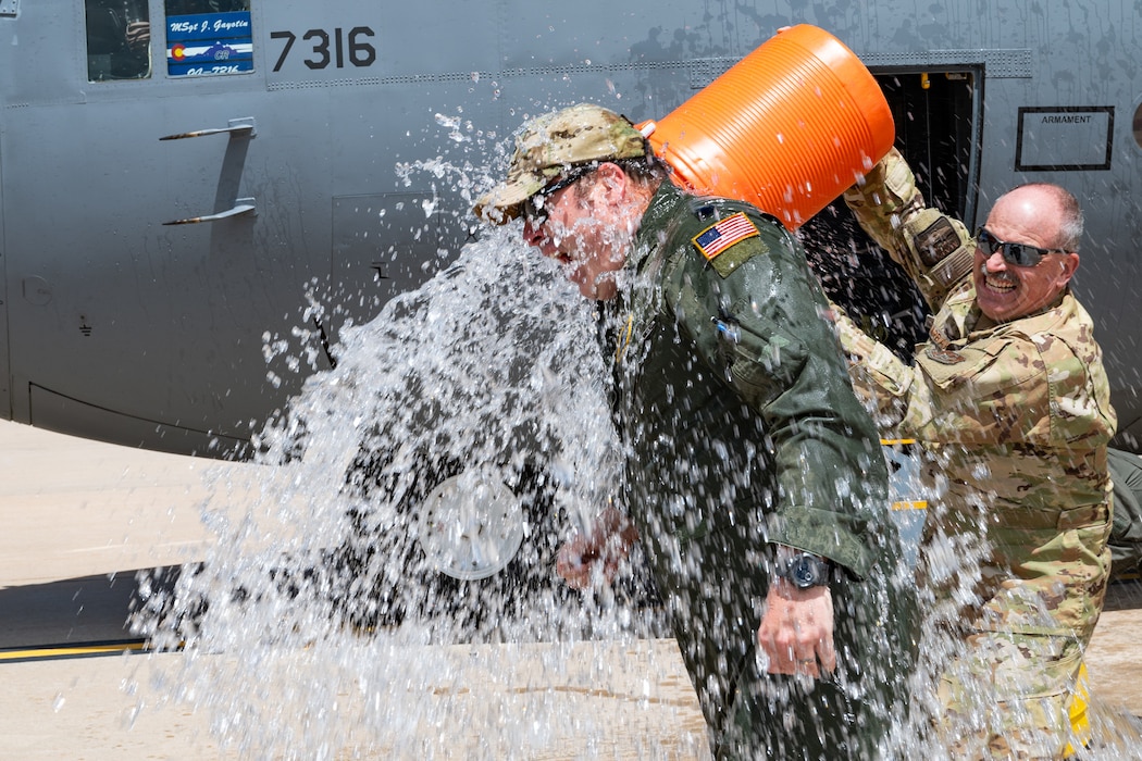 A man in uniform dumping a container of water onto another man in a flight suit with a military aircraft in the background.