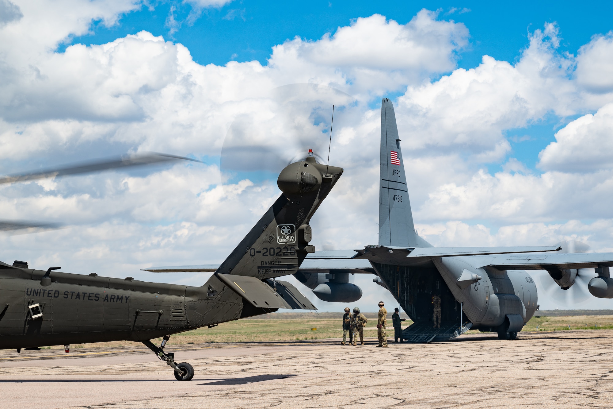 A helicopter and fixed-wing aircraft parked on a flight line with military personnel at the back of the aircraft.
