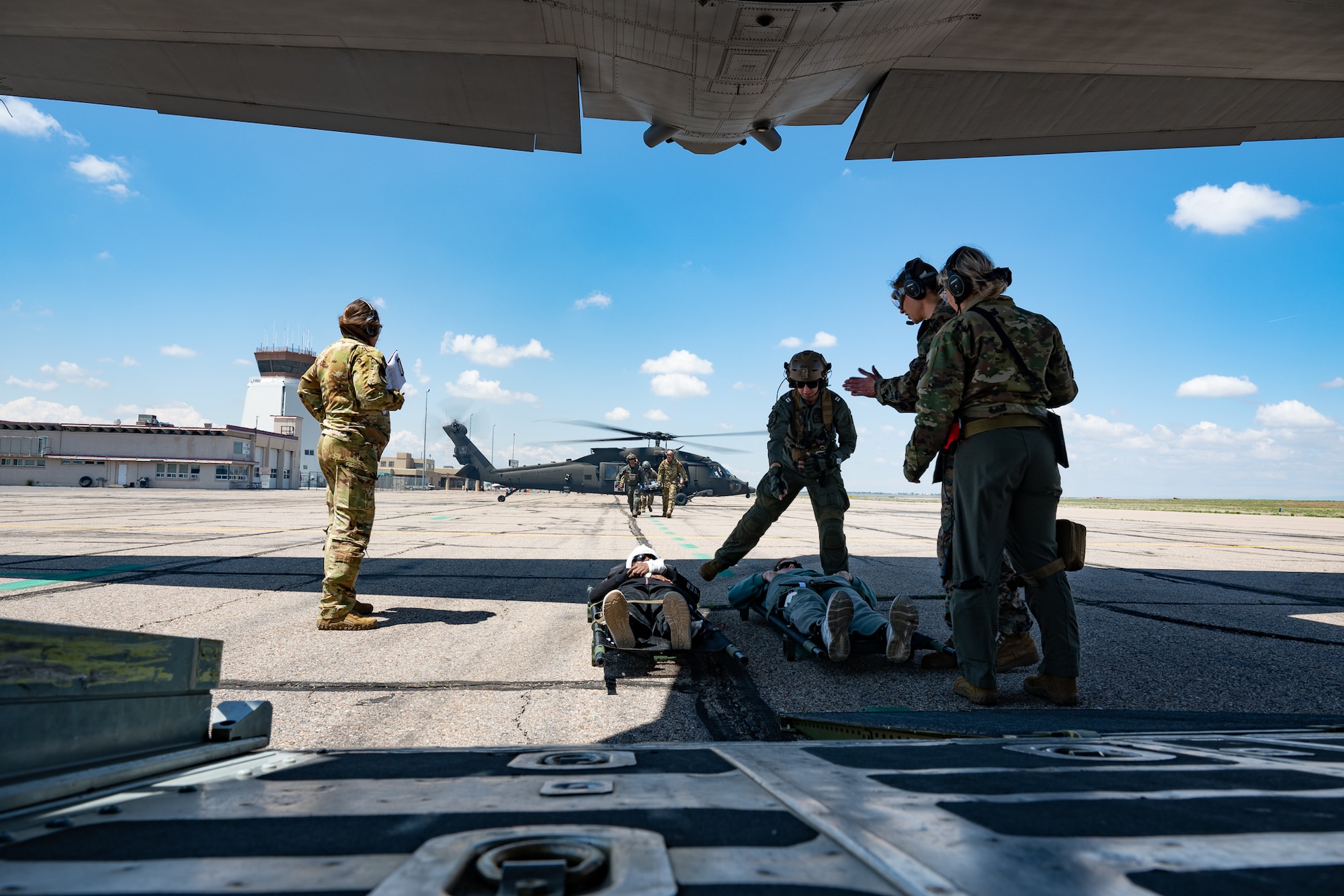 A group of servicemembers in front of litter patients behind the cargo bay of a military aircraft in the foreground with a helicopter in the background and a separate team approaching.