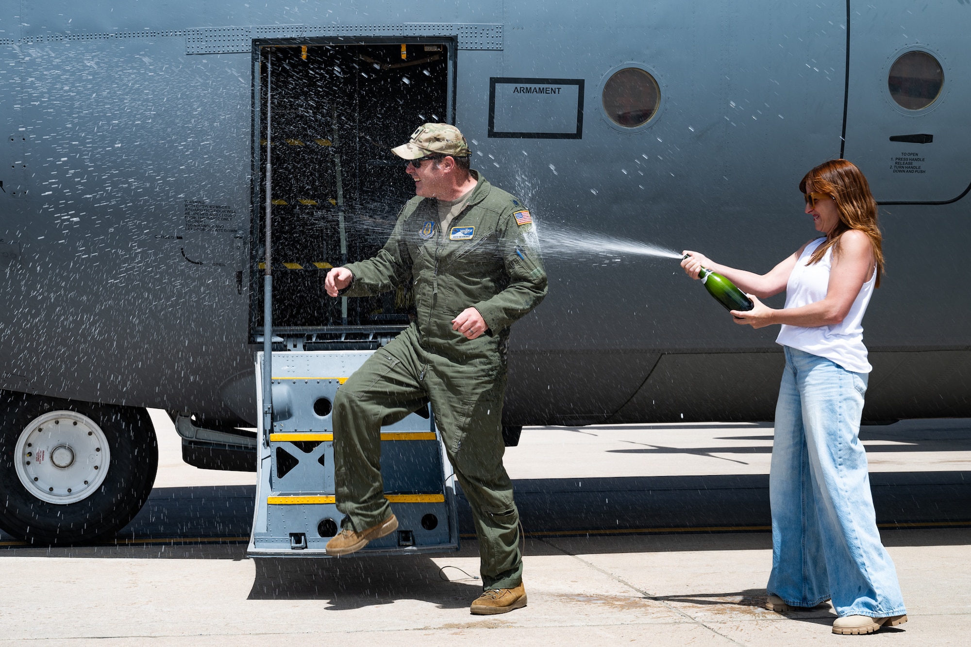 A man in a flight suit is sprayed with champagne by his wife in front of a military aircraft.