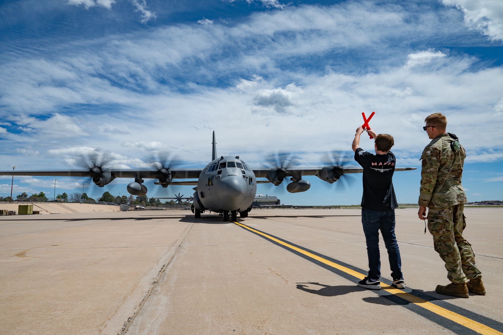 A child in civilian clothes raises marshalling wands in the air while a service member stands next to him and a military aircraft approaches them.