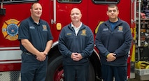 Three people wearing Tobyhanna Army Depot Fire & Emergency Services apparel stand in front of a firetruck and pose for a photo.