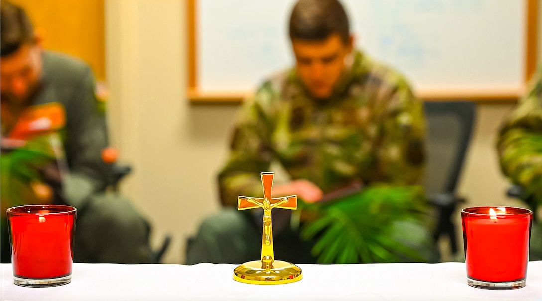 A person in a military uniform bows there head in prayer in the background. in the foreground are two red candles burning with a gold cross on the table between them.