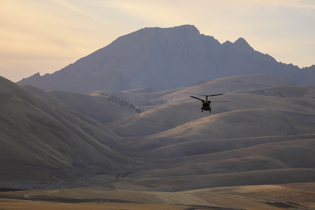 A helicopter flies above brown, rolling mountains at sunset.