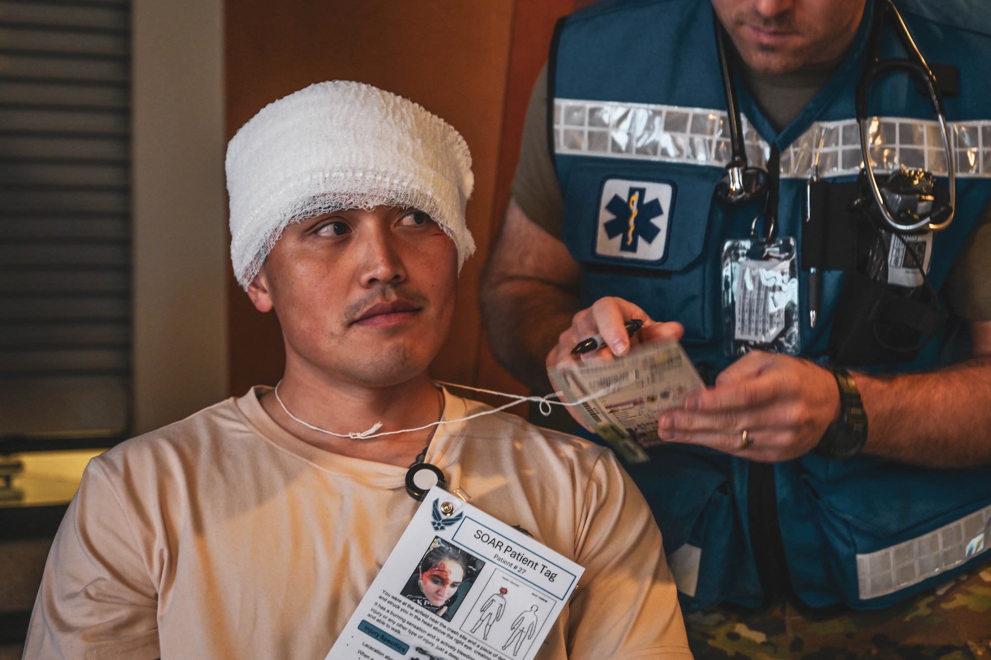 A simulated patient with a head bandage is cared for by a man in uniform.