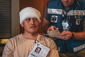 A simulated patient with a head bandage is cared for by a man in uniform.