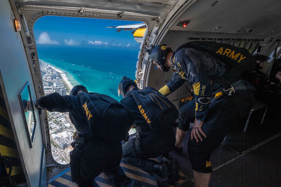 Three soldiers in flight gear lean out of an airplane above a city and ocean.
