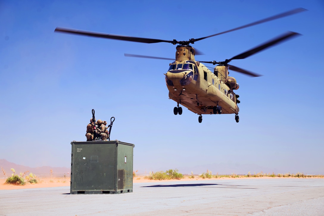 Three soldiers kneeling on top of a container prepare to hook it to a descending helicopter on a runway.