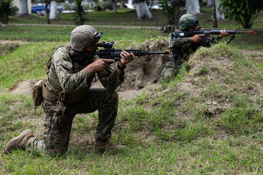 A U.S. service member and Guatemalan service member kneel on the grass and aim their guns