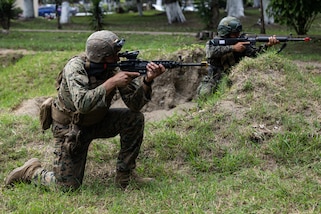 A U.S. service member and Guatemalan service member kneel on the grass and aim their guns