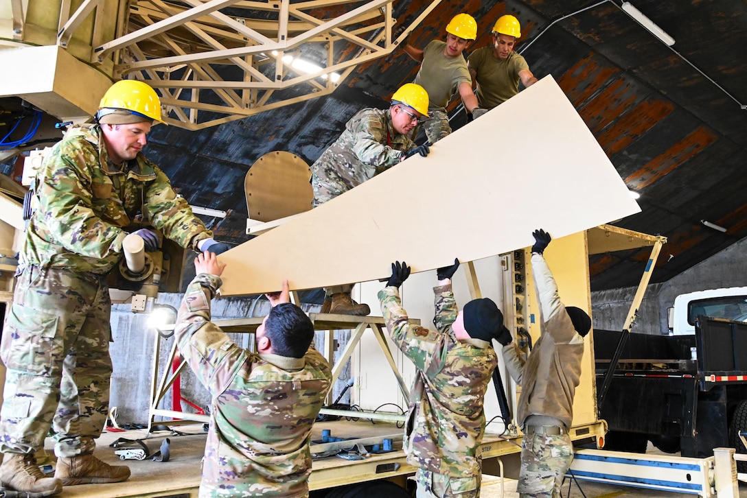 Seven airmen, four of them wearing construction helmets, hold up a large piece of wood inside a facility.