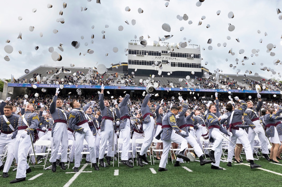 U.S. Military Academy graduates in dress uniforms throw their caps toward the sky during a ceremony at a football stadium.