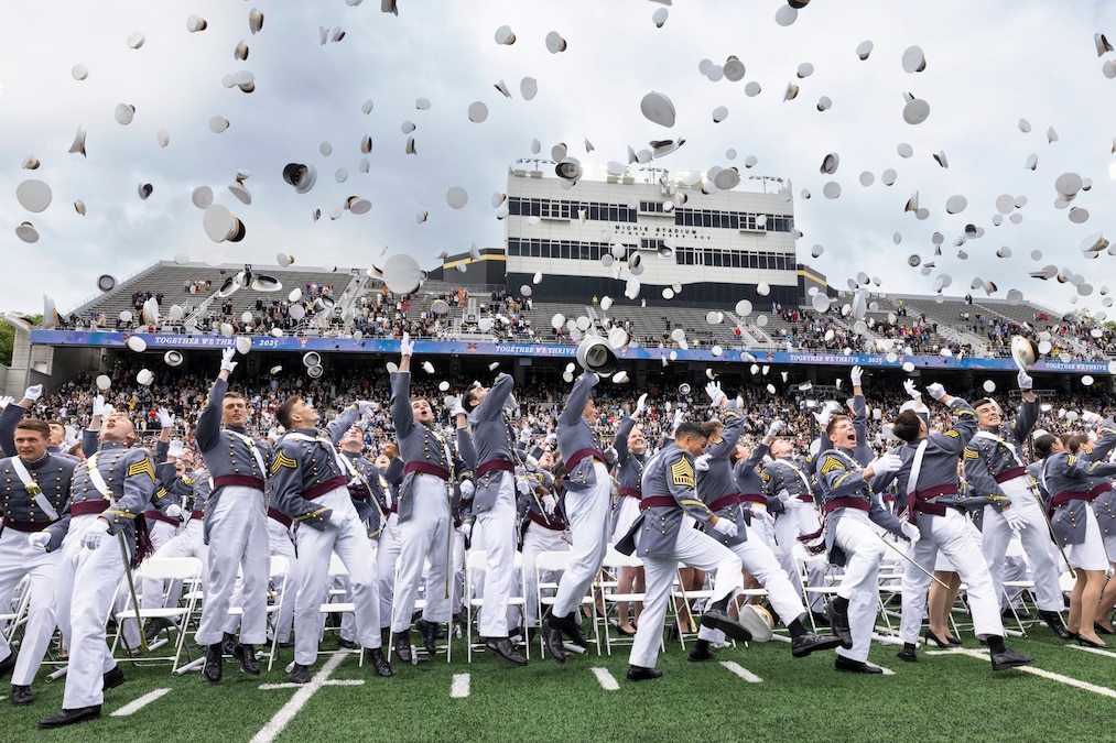 U.S. Military Academy graduates in dress uniforms throw their caps toward the sky during a ceremony at a football stadium.