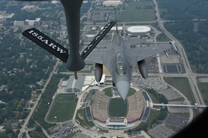A refueling boom hangs over an F-16 fighter jet as it flies over Jack Trice Stadium