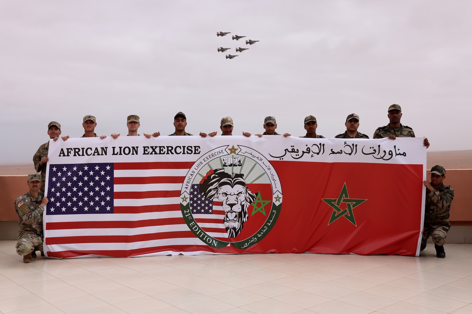 U.S. Marines and Soldiers from the Royal Moroccan Armed Forces pose for a group photo while F16 fighter jets pass overhead at Cap Draa during African Lion 25 (AL25) in Tan-Tan, Morocco, May 23, 2025. AL25, the largest annual military exercise in Africa, brings together over 50 nations, including seven NATO allies and 10,000 troops to conduct realistic, dynamic and collaborative training in an austere environment that intersects multiple geographic and functional combatant commands. Led by U.S. Army Southern European Task Force, Africa (SETAF-AF) on behalf of the U.S. Africa Command, AL25 takes place from April 14 to May 23, 2025, across Ghana, Morocco, Senegal, and Tunisia. This large-scale exercise will enhance our ability to work together in complex, multi-domain operations—preparing forces to deploy, fight and win.