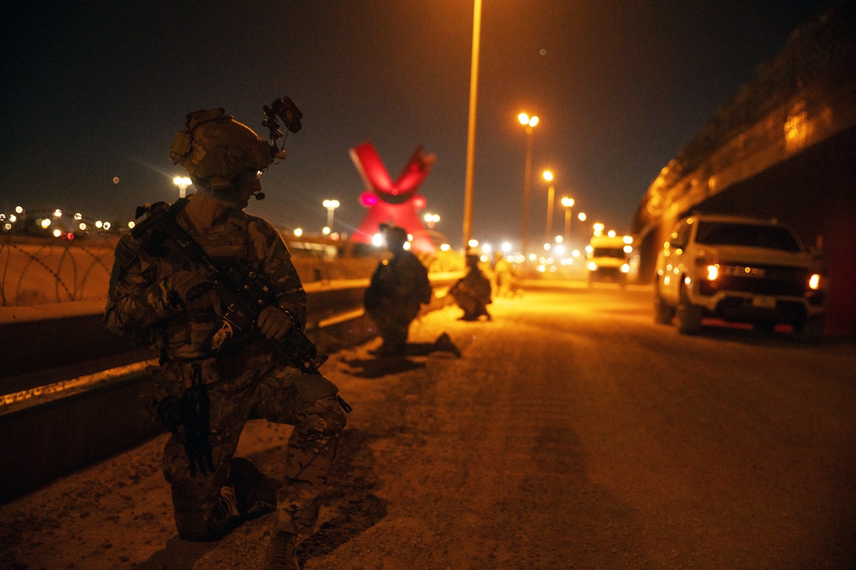 Soldiers in combat gear kneel on a road with streetlights, two trucks and other objects in the background at night.