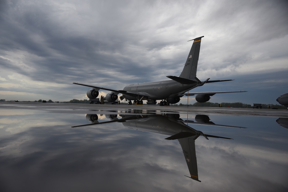 A KC-135 air refueling plane is still on a flight ramp. Its reflection shows in water beneath it.