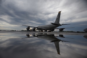 A KC-135 air refueling plane is still on a flight ramp. Its reflection shows in water beneath it.