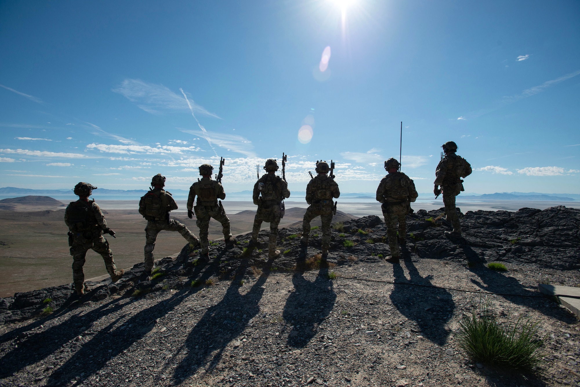 Soldiers, shown from behind in silhouette, look over arid, hilly terrain against a sunny, blue sky.