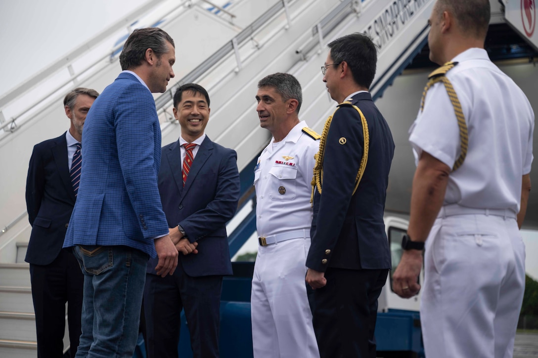 Defense Secretary Pete Hegseth speaks with military personnel and civilians outside the steps of an aircraft.