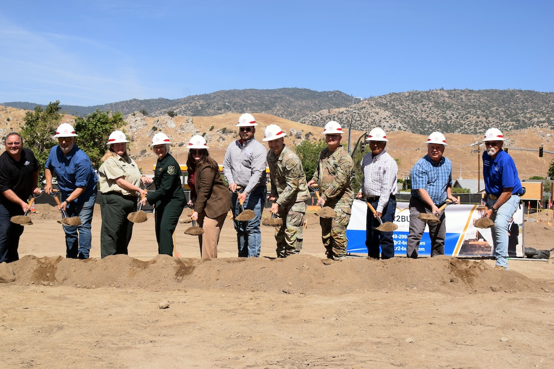 A mix of people wearing hard hats hold up shovels of dirt in front of golden foothills.