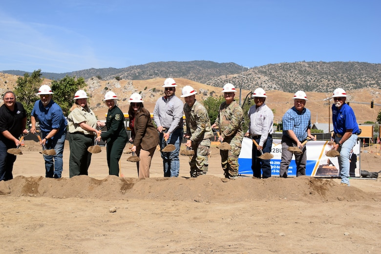 A mix of people wearing hard hats hold up shovels of dirt in front of golden foothills.