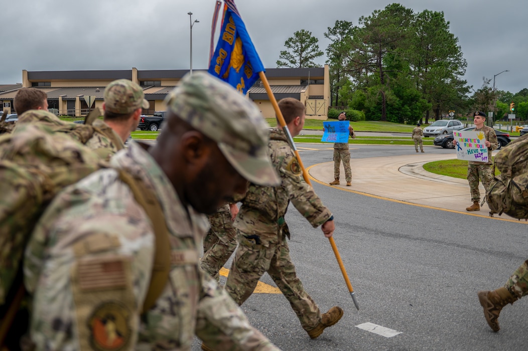 A man holding a sign is seen in the background, as a group march past.