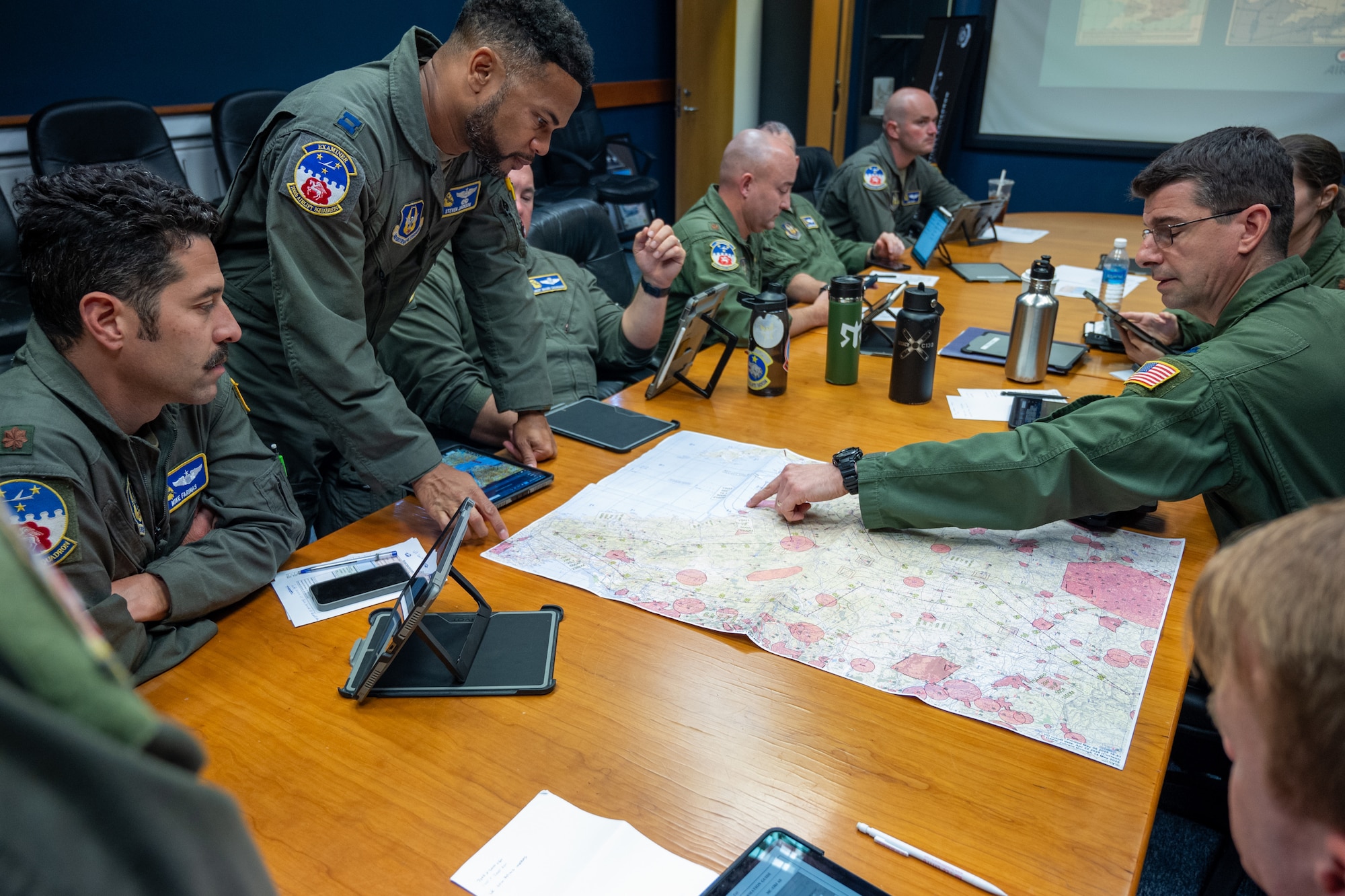 Airmen in flight suits sit around a table and point to a map