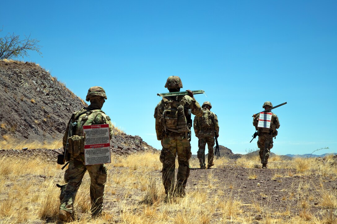 Four soldiers in combat gear walk in desert-like conditions with a hill next to them on a sunny day.