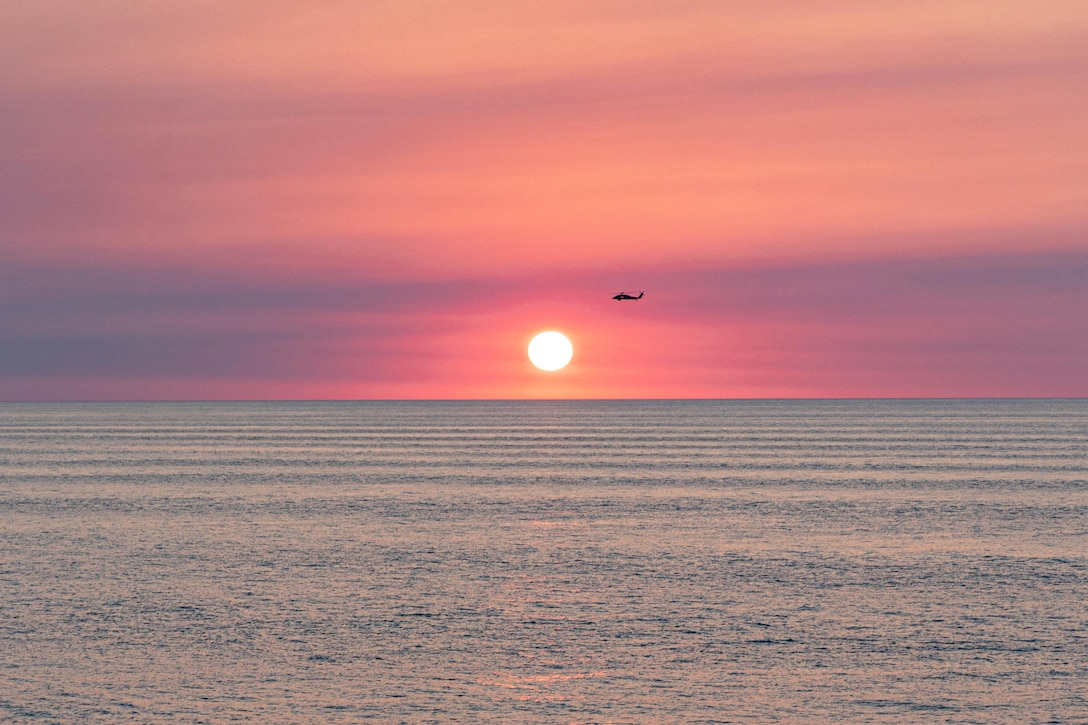 A bright sun shines on the horizon as a helicopter in silhouette flies in a pinkish, blue sky over a body of water.
