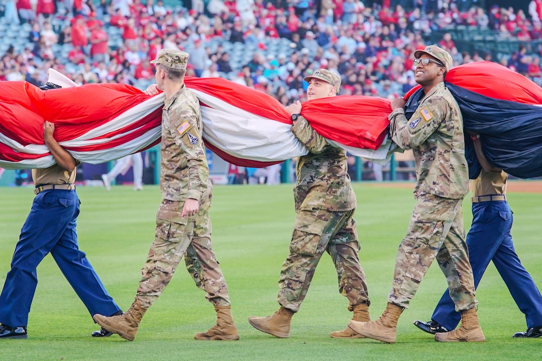 Guardians and fellow service members carry a rolled up American flag across a baseball field as fans sit the bleachers of the stadium.
