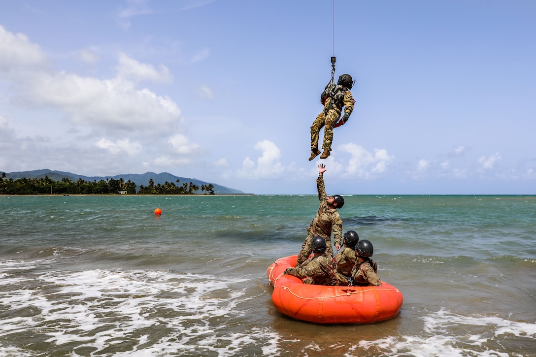 A soldier in a harness is lifted above four soldiers in an orange raft on the ocean.
