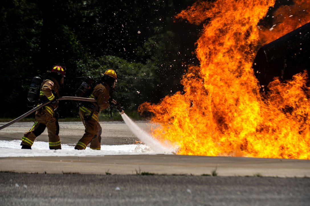 Two Air Force firefighters in suits use a hose on a simulated fire.
