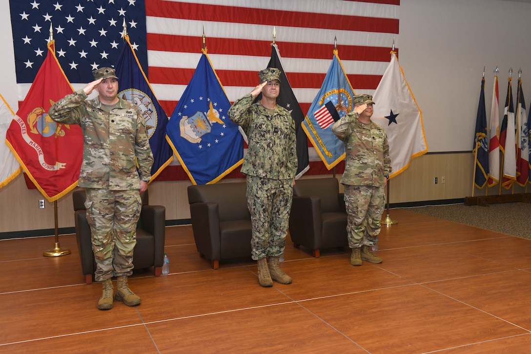 Three men wearing camouflage military uniforms stand on a stage saluting with U.S. flag and U.S. military branch flags behind them.