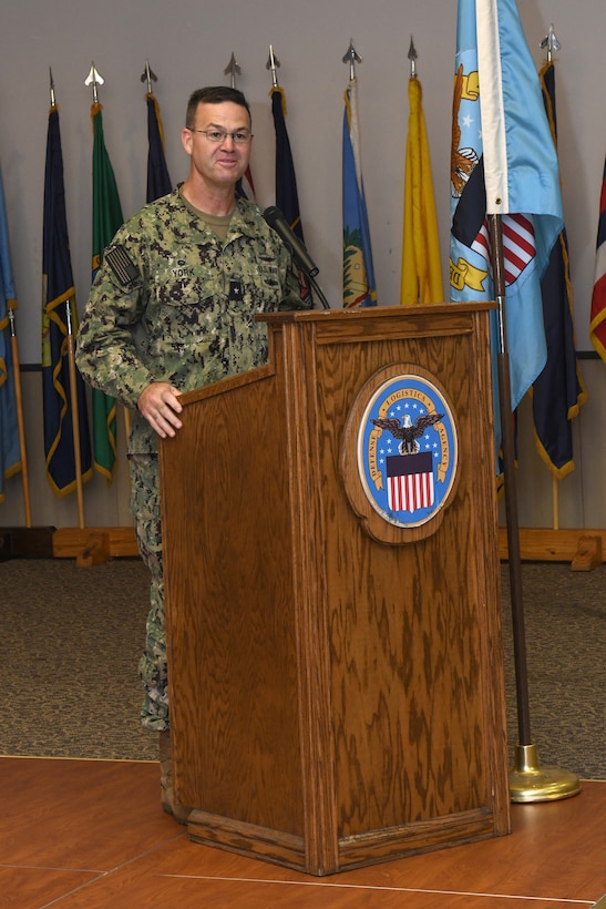 A man wearing a camouflage military uniform stands on a stage behind a lectern giving a speech.