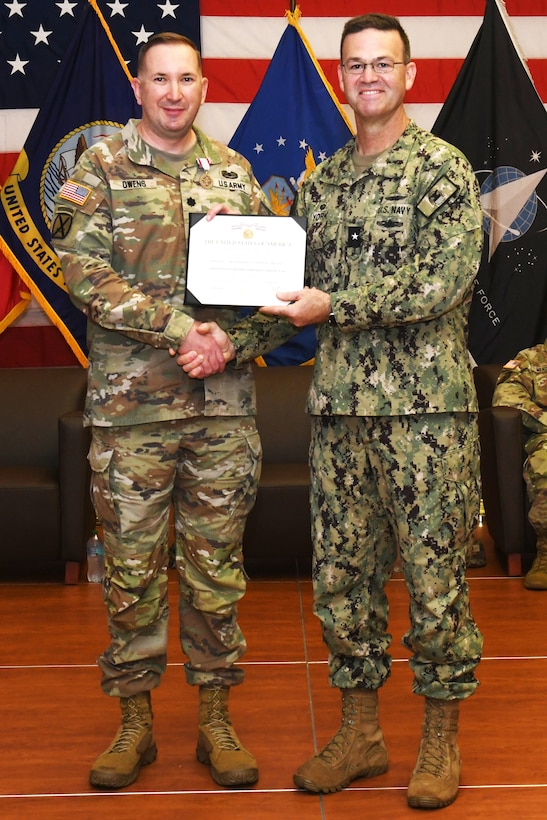 Two men wearing camouflage military uniforms stand on a stage holding a certificate and shaking hands.